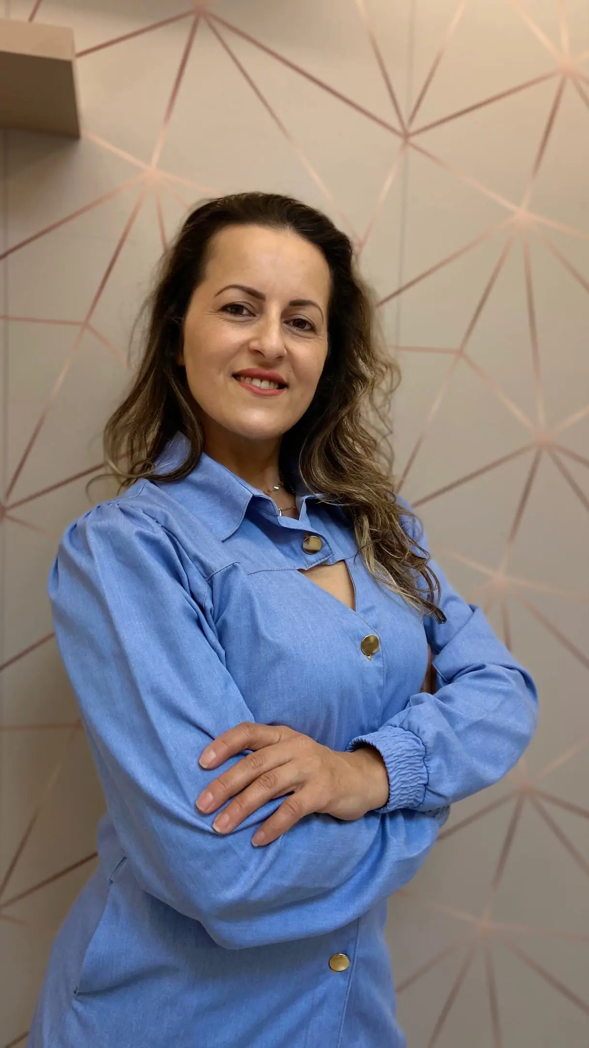 Andreia Firmino, esteticista em Joinville, sorrindo de braços cruzados em frente a uma parede rosé